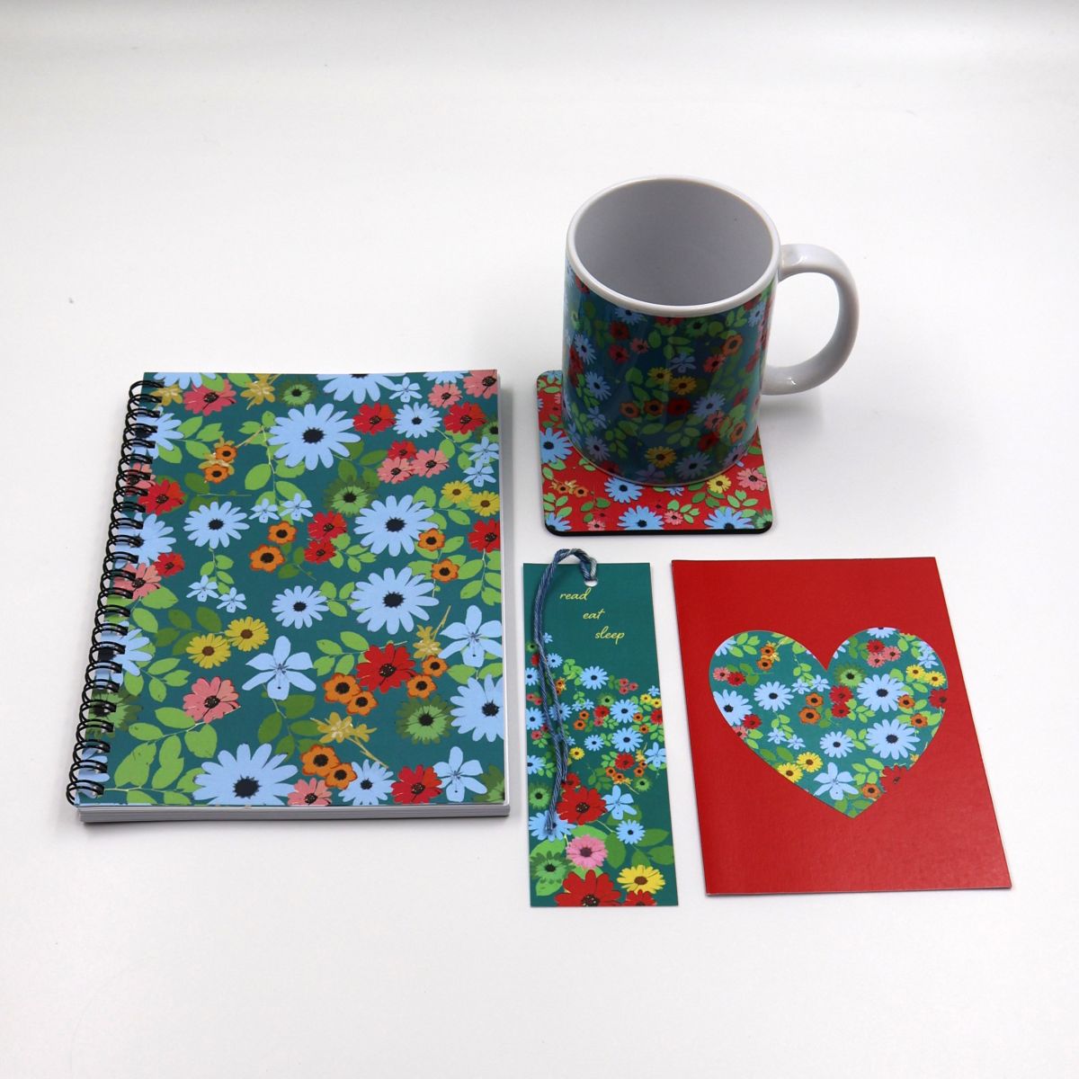 Individual lifestyle stationery items shown separately flat lay style including a thick paper A5 notebook a pattern printed ceramic mug a square desk coaster a bookmark and card