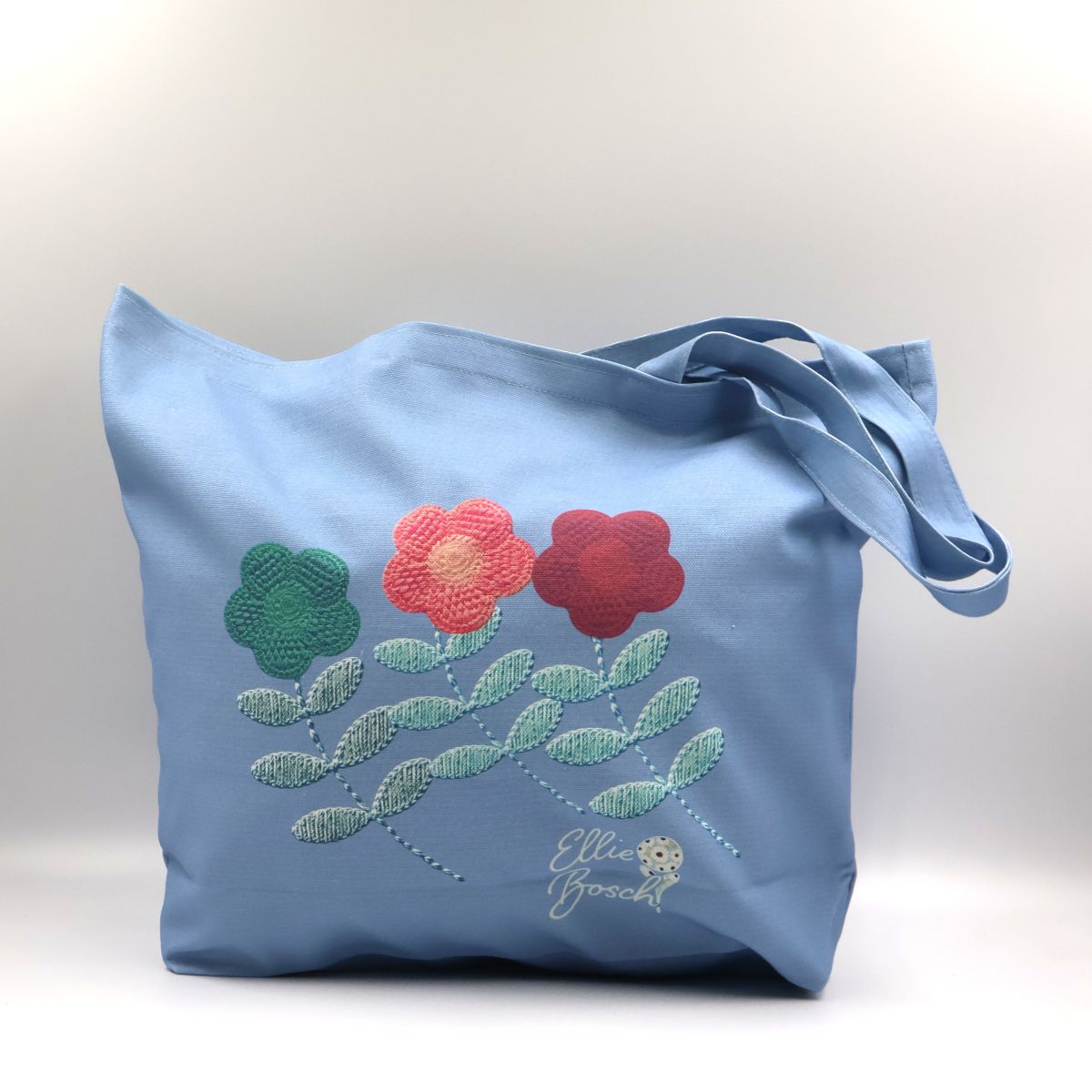 Handmade South African light denim blue cotton canvas tote bag showing the front panel printed with orange red and green crochet daisy flower motifs