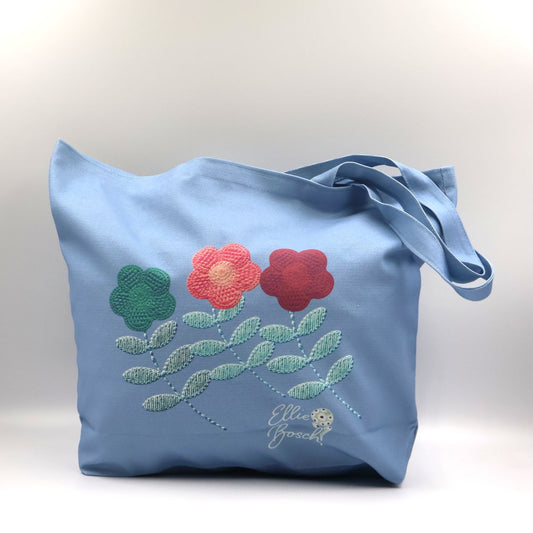 Handmade South African light denim blue cotton canvas tote bag showing the front panel printed with orange red and green crochet daisy flower motifs