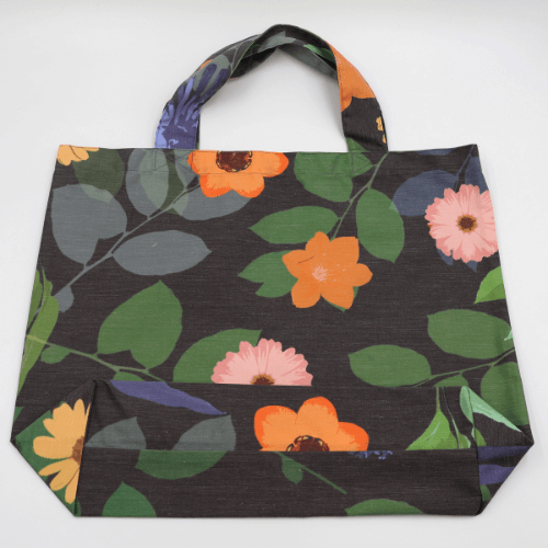 Flat view of a big, durable tote bag with a bright orange and yellow daisy design on dark grey background.